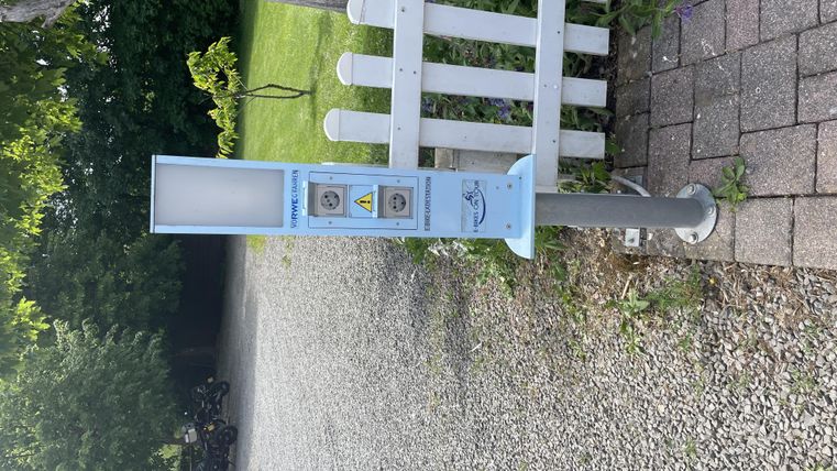 Eine öffentliche Ladestation für Elektrofahrzeuge steht auf einem gepflasterten Weg. Im Hintergrund sind Bäume und ein Zaun zu sehen.