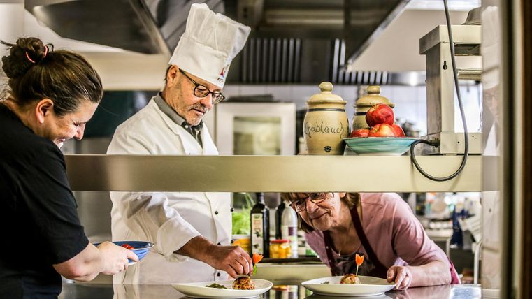 Eine Küche mit einem Chefkoch, der gerade ein Gericht anrichtet. Zwei weitere Personen beobachten und helfen dabei.