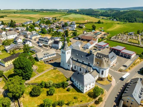 Eine malerische Dorfansicht mit mehreren Gebäuden und einer Kirche. Im Hintergrund sind grüne Felder und sanfte Hügel zu sehen.