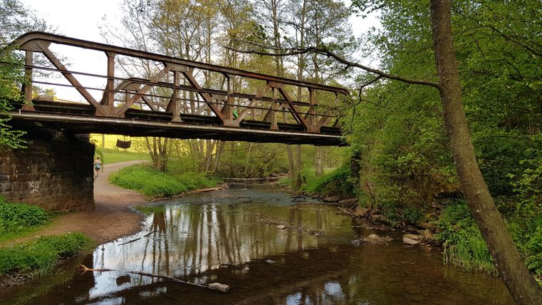 An old wooden bridge over a small river. Green trees and bushes surround the scene.