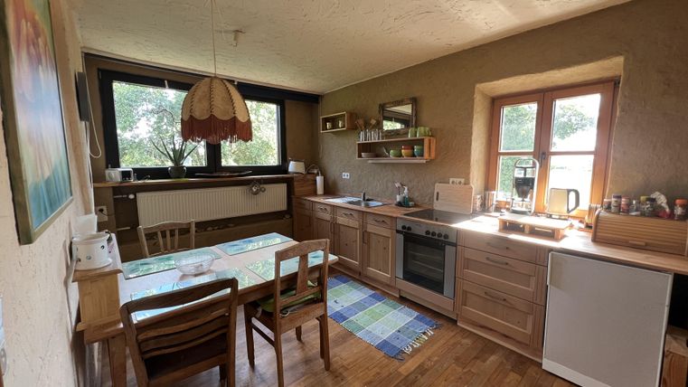 Eine gemütliche Küche mit Holzschränken und einem Esstisch. Durch große Fenster fällt viel Licht in den Raum.