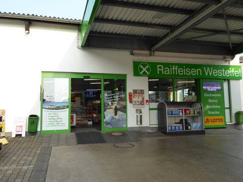 Ein Markt mit einem Eingangsbereich in grünen Farben. Es sind Werbetafeln und Regale sichtbar, die verschiedene Produkte anzeigen.
