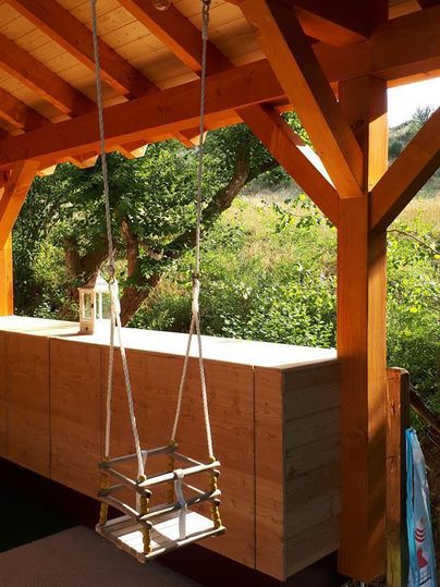 Eine Holzschaukel hängt unter einem überdachten Bereich. Im Hintergrund sind grüne Pflanzen und ein sonniger Himmel sichtbar.