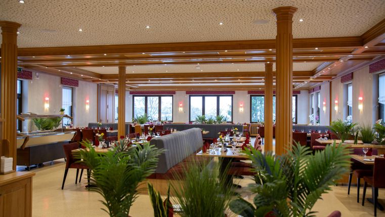 Ein elegantes Restaurant mit stilvollen Tischen und Pflanzen. Große Fenster sorgen für viel Tageslicht und eine einladende Atmosphäre.