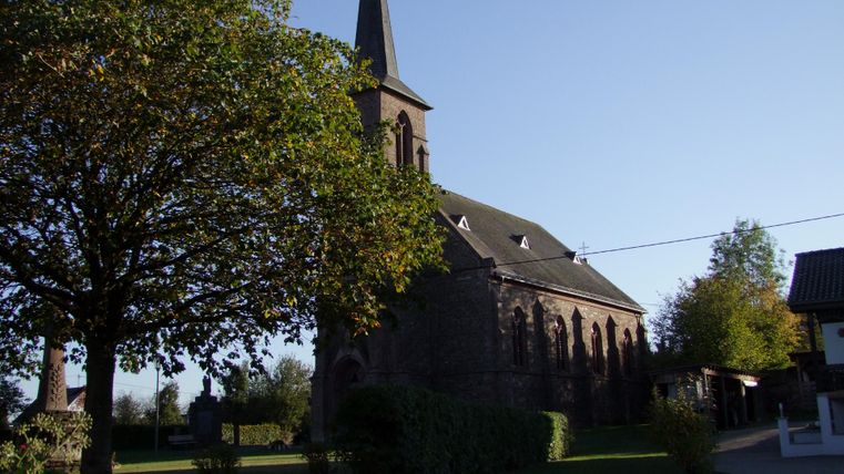 Een oude kerk met een hoge toren staat naast een grote boom. De lucht is helder en blauw, wat de rustige sfeer versterkt.