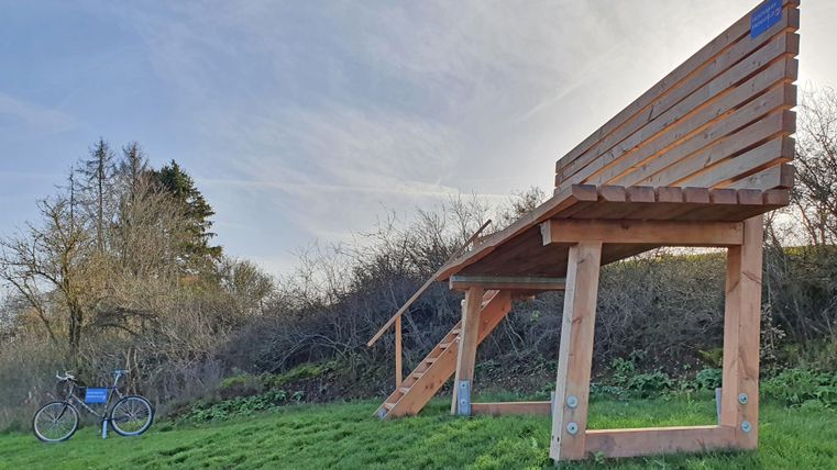 Eine große Holzbank steht auf einer Wiese, daneben ein Fahrrad. Im Hintergrund sind Bäume und ein klarer Himmel zu sehen.
