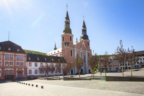 Ein historisches Gebäude mit zwei Türmen steht in einem weitläufigen Platz. Umgeben von Bäumen und bei klarem blauen Himmel.
