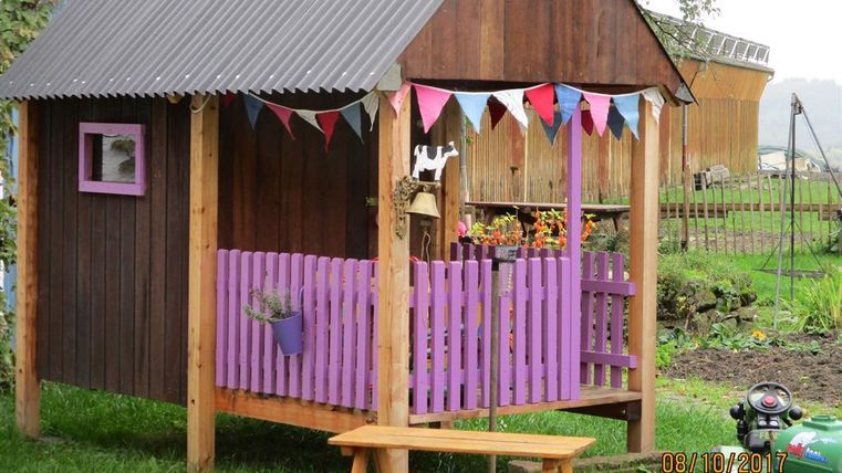 Ein kleines, buntes Spielhaus mit einer Holzveranda und einem Zaun in Lila. Vor dem Haus steht ein Holztisch mit Bänken.