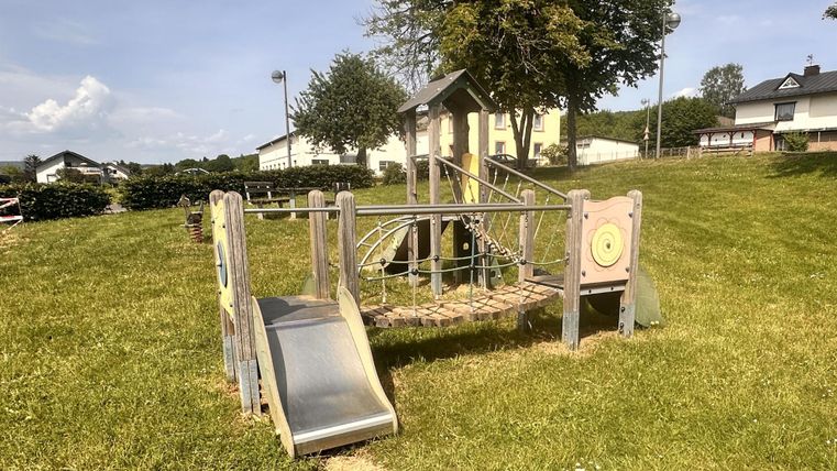 Ein Spielplatz mit einer Rutsche und einem Klettergerüst auf einer grünen Wiese. Im Hintergrund sind einige Bäume und Häuser zu sehen.