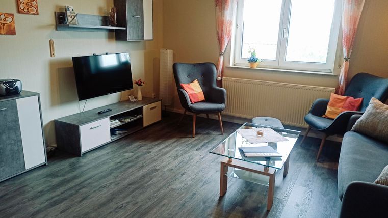 Ein modernes Wohnzimmer mit einem Fernseher und einer gemütlichen Sitzecke. Große Fenster lassen viel Licht herein und es gibt einige dekorative Elemente.