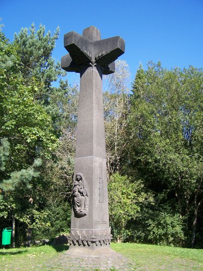 Ein hohes Monument in Form eines Kreuzes steht in einer grünen Umgebung. Die Statue zeigt eine figurative Darstellung an der Basis des Gedenksteins.