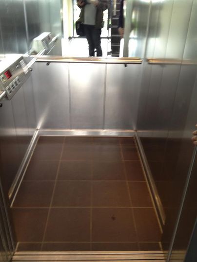 Ein leerer Aufzug mit metallischen Wänden und einem dunklen Boden. Im Hintergrund ist eine reflektierende Oberfläche sichtbar.