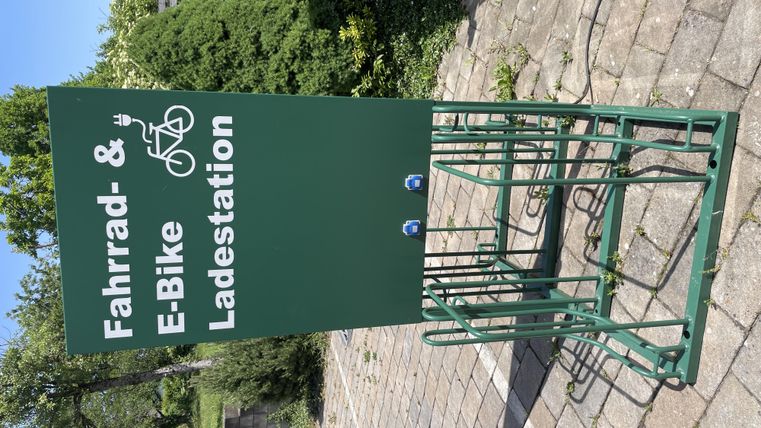 A bicycle and e-bike charging station is located on a paved area. In the background, trees and shrubs are visible.
