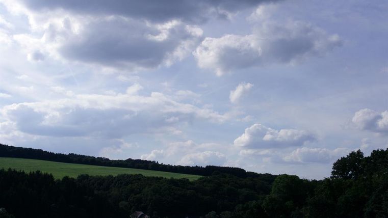 Ein Himmel mit vielen Wolken über einer grünen Landschaft. Im Hintergrund sind Wälder und einige Gebäude sichtbar.