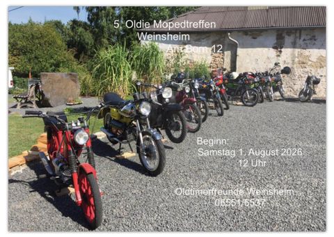 A row of vintage mopeds is lined up on a gravel area. In the background, green vegetation and a building can be seen.