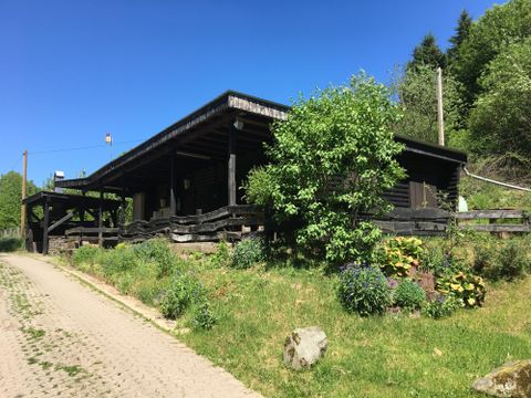 Ein Holzhaus umgeben von grünen Bäumen und Blumen. Der Himmel ist klar und blau.