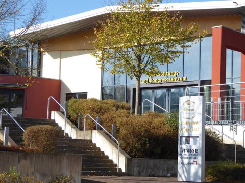 Ein modernes Veranstaltungs- und Kongresszentrum mit einer großen Fensterfront. Vor dem Gebäude befinden sich Treppen und eine gepflegte Grünanlage.