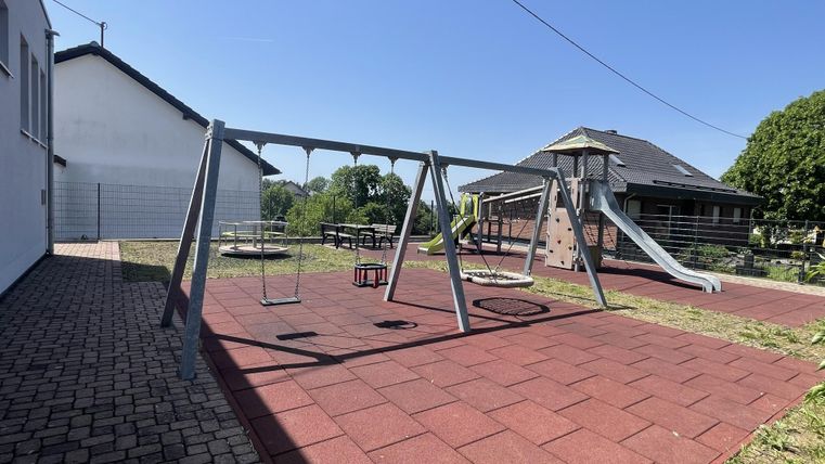 Ein Spielplatz mit einer Schaukel und einer Rutsche. Der Boden ist mit roten Gummiplatten ausgelegt und es gibt viel Grasflächen.