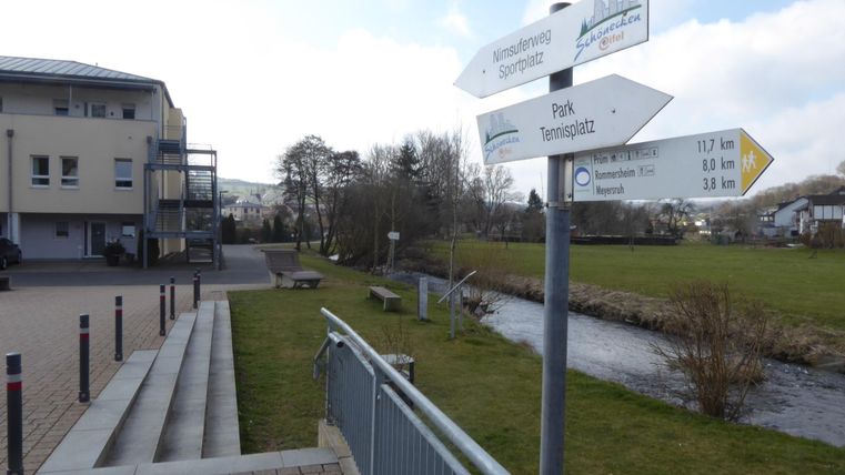 Ein Wegweiser zeigt verschiedene Ziele an, darunter einen Park und den Stadtplatz. Im Hintergrund sind ein Gebäude und eine grüne Wiese zu sehen.