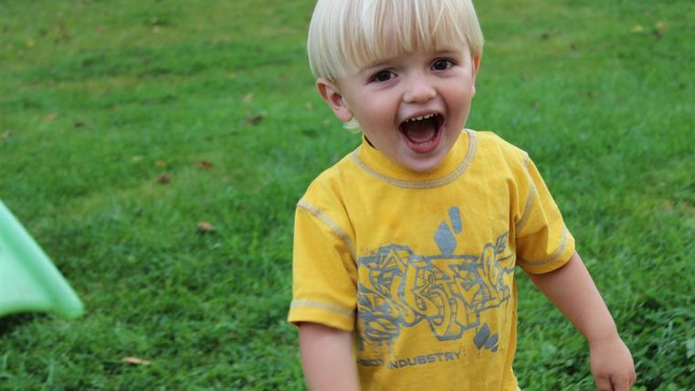 Ein kleiner Junge mit blonden Haaren trägt ein gelbes T-Shirt. Er lacht und spielt auf einer grünen Wiese.