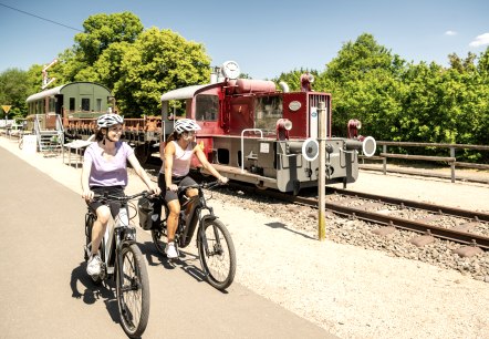 Zwei Radfahrerinnen auf einem Radweg neben einer alten Lokomotive im Eisenbahnmuseum Pronsfeld, umgeben von grünen Bäumen., © Eifel Tourismus GmbH, Dominik Ketz