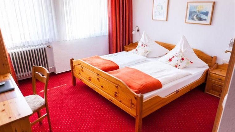 Ein gemütliches Schlafzimmer mit einem großen Holzbett und orangefarbenen Kissen. Die Wände sind hell, und es gibt einen roten Teppich sowie große Fenster, die viel Licht hereinzulassen.