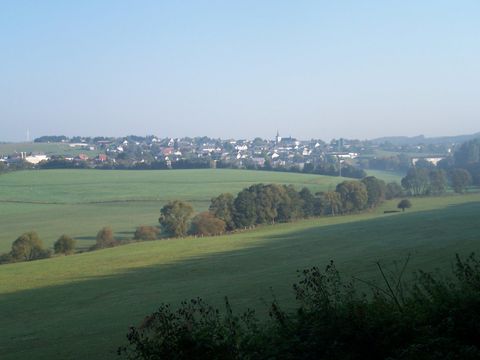 Eine malerische Landschaft mit sanften Hügeln und einem kleinen Dorf im Hintergrund. Der Himmel ist klar und sonnig.