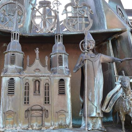 Eine kunstvolle Metallskulptur zeigt einen Bischof mit einem Stab und eine Kirche. Im Hintergrund sind weitere Verzierungen zu sehen, die an Geschichten oder Traditionen erinnern.