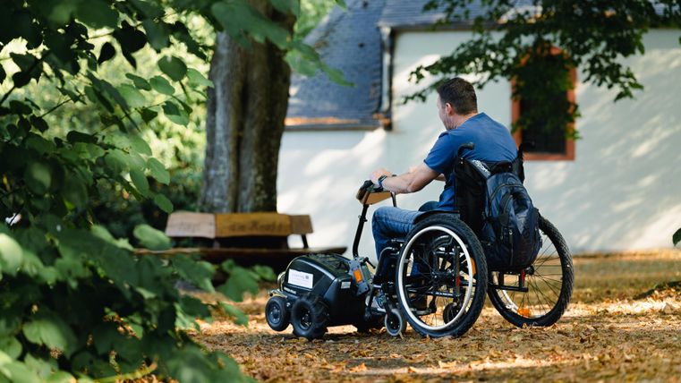 Un homme est assis dans un fauteuil roulant et observe les environs dans une zone semblable à un parc. Le sol est couvert de feuilles d'automne, et en arrière-plan, un bâtiment est visible.