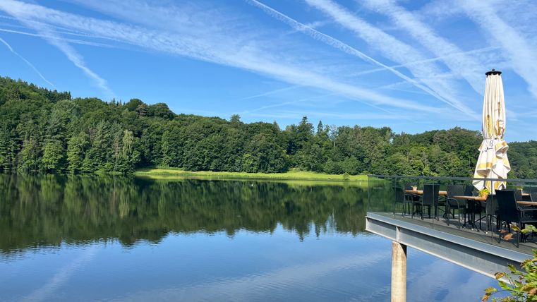 Lac de barrage de Bitburg avec terrasse et parasol, entouré de forêt et d'un ciel bleu avec des traînées de condensation.