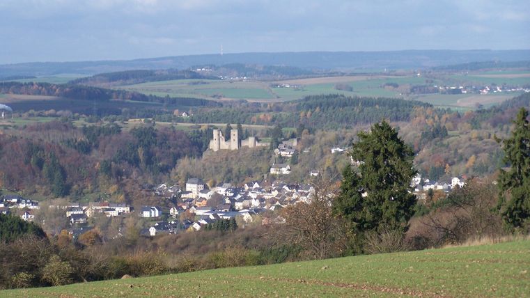 Eine malerische Landschaft mit sanften Hügeln und einem kleinen Dorf. Im Hintergrund sind alte Burgruinen zu sehen.