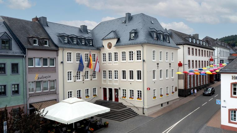 Ein historisches Gebäude mit mehreren Fahnen vor der Fassade. Auf der Straße sind bunte Wimpel und ein Restaurant mit Sonnenschirm zu sehen.