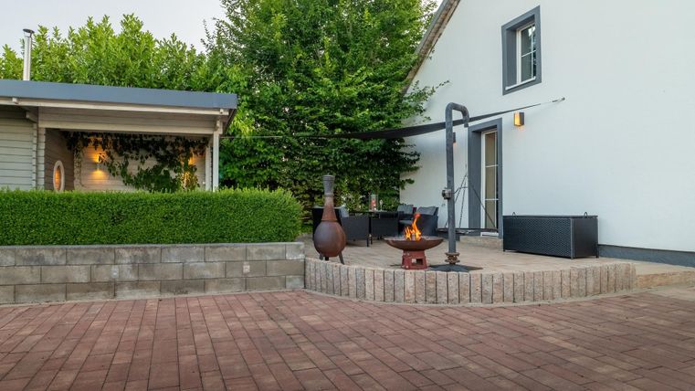 Ein gemütlicher Außenbereich mit einer gepflasterten Terrasse und einer Feuerstelle. Im Hintergrund stehen ein paar Gräser und ein modernes Gebäude.