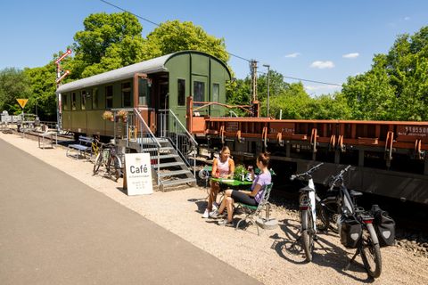 Eine gemütliche Cafészene neben einem ehemaligen Zugwagen. Zwei Personen sitzen draußen und genießen ihren Aufenthalt, während Fahrräder in der Nähe abgestellt sind.