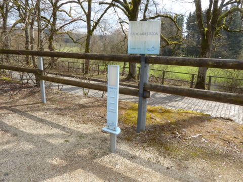 Eine Hinweistafel steht an einem Weg mit einer Skala daneben. Der Bereich ist von Bäumen umgeben und hat einen klaren Blick auf die Landschaft.