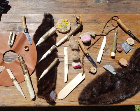 Eine Vielzahl von handgefertigten Werkzeugen und Materialien liegt auf einem Tisch. Darunter sind Holzstücke, Steinwerkzeuge und ein Lederstück zu sehen.