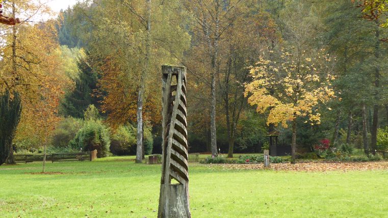 Eine kunstvolle Holzskulptur steht im Grünen in einem parkähnlichen Bereich. Bunte Bäume umgeben die Skulptur und verleihen der Szene eine herbstliche Atmosphäre.