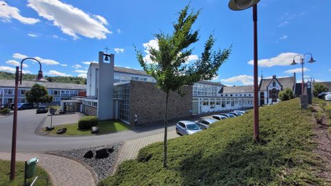Een modern gebouw in een rustige omgeving met bomen en een heldere blauwe lucht. Parkeerplaatsen zijn dichtbij en het landschap is goed onderhouden.