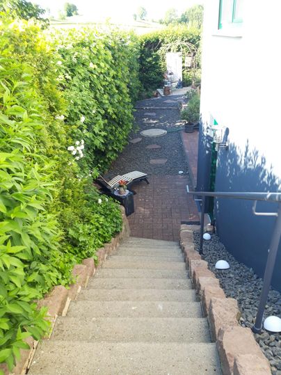 Ein geschützter Gartenweg mit Stufen, umgeben von üppigem Grün. Der Weg führt zu einer gepflasterten Fläche mit Sitzgelegenheiten.