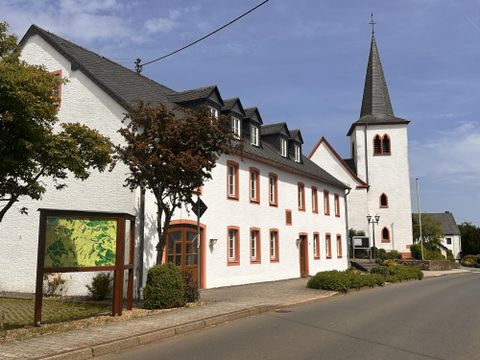 Ein charmantes Gebäude mit weißer Fassade und roten Akzenten steht neben einer Kirche mit spitzem Turm. Die Straße verläuft ruhig und ist von Bäumen und Sträuchern gesäumt.