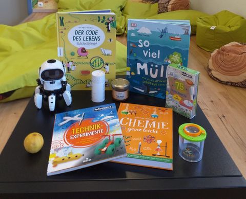Ein Tisch mit verschiedenen Büchern, darunter Titel über Technik und Chemie. Außerdem sind ein kleiner Roboter, ein Glas und ein Apfel zu sehen.