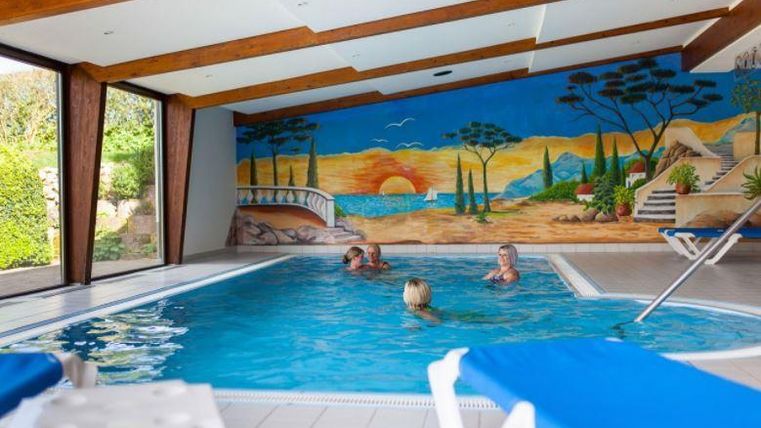 Ein Innenpool mit einem bunten Wandgemälde und mehreren Personen, die schwimmen. Es gibt Liegen und viel natürliches Licht.