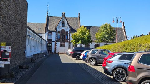 Une étroite rue avec des voitures garées mène à un bâtiment historique. Le temps est ensoleillé et le ciel est bleu.