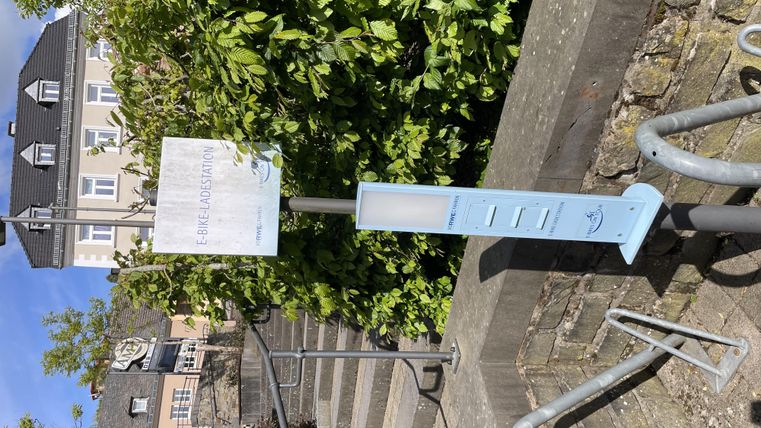 Eine Informationssäule steht an einer Treppe, umgeben von grünen Pflanzen. Im Hintergrund sind einige Häuser zu sehen.