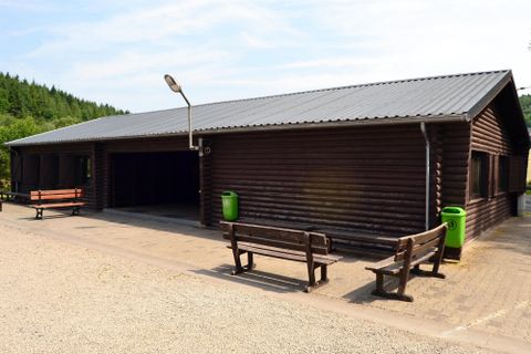 Ein schlichtes Holzgebäude mit einem grauen Dach. Vor dem Gebäude stehen mehrere Bänke und es gibt eine grüne Abfalltonne.