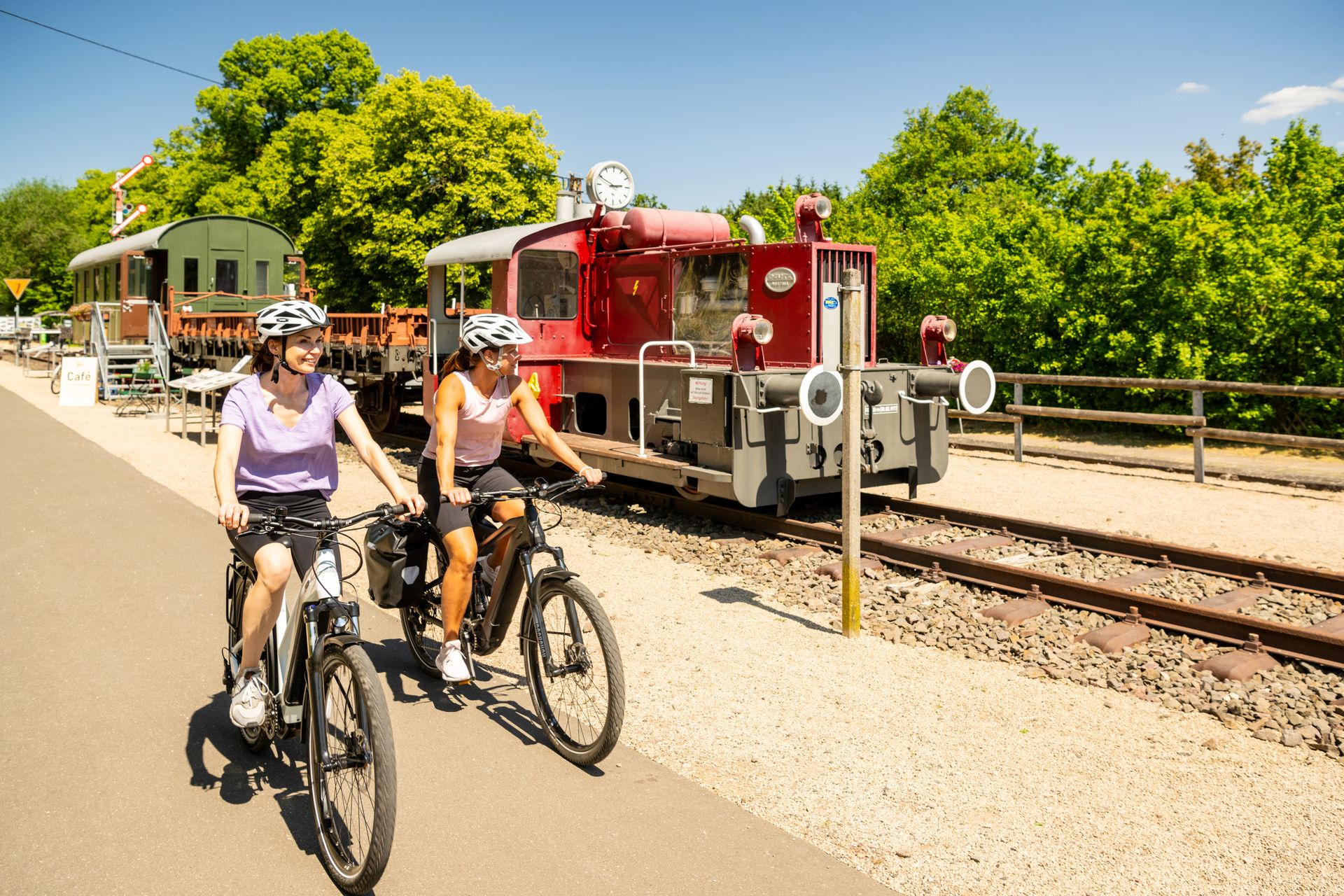 Zwei Radfahrerinnen fahren auf einem Radweg an einer alten Lokomotive vorbei.