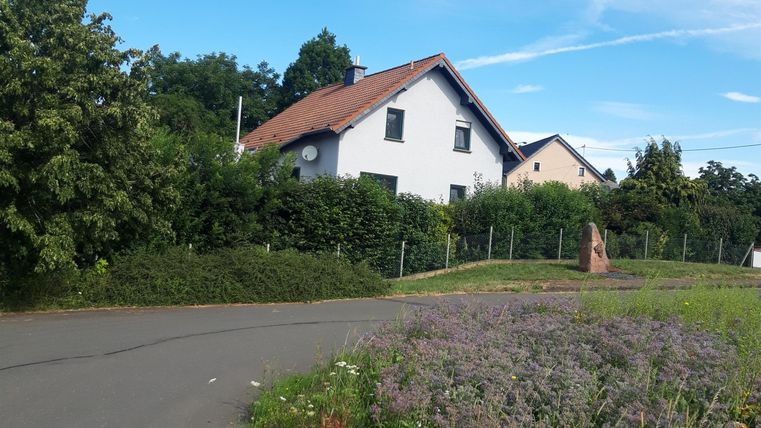 Ein ruhiges Wohngebiet mit einem weißen Haus und einem roten Dach. Umgeben von grünen Bäumen und blühenden Pflanzen unter einem blauen Himmel.