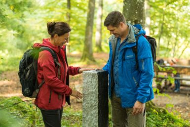 Zwei Personen stehen im Wald neben einem schwarz-weißen Grenzstein.