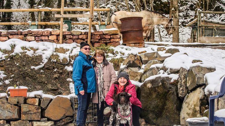 Eine Gruppe von drei Personen steht in einer verschneiten Landschaft und posiert mit einem Hund. Im Hintergrund sind einige Bäume und ein Holzstapel zu sehen.