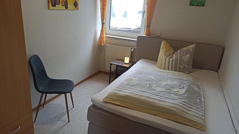 Ein einfaches Schlafzimmer mit einem Einzelbett, einem Stuhl und einem kleinen Tisch. Es gibt Fenster mit Gardinen und Wände, die mit Kunstwerken dekoriert sind.